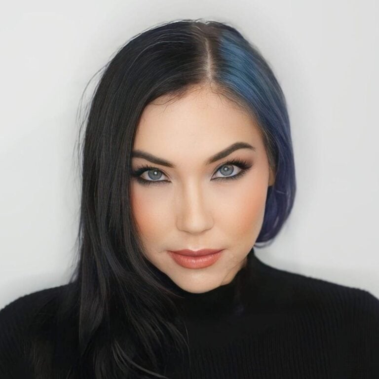 Close-up portrait of a woman with striking blue highlights in her dark hair, intense blue eyes, and natural makeup, set against a plain white background.
